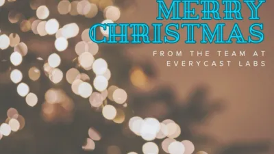 The Everycast Labs logo (three labradors in a circle, one wearing a headset) sits in the top right-hand corner of the image. Below, blue and white writing reads "Merry Christmas from the team at Everycast Labs". The background is bokeh effect Christmas lights on a tree. There is a festive arrow in the bottom right-hand corner pointing to the next image.